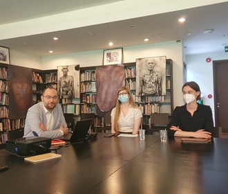 Jeden mężczyzna oraz dwie kobiety siedzą przy stole w pomieszczeniu z regałami, na których znajdują się książki/One man and two women are sitting at the table in the room with bookshelves on which there are books